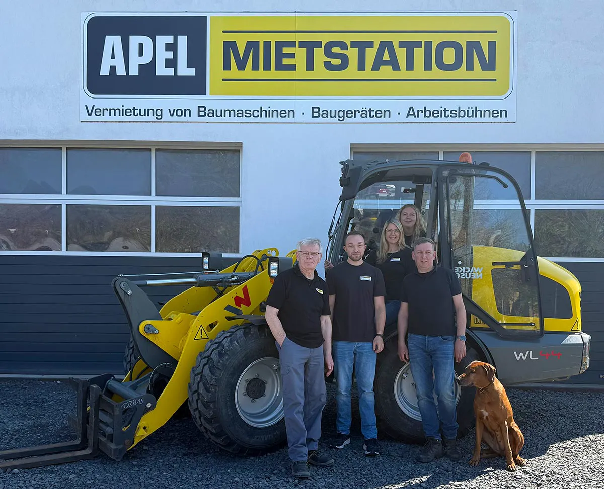 Fünf Personen und ein Hund stehen vor einem gelben Baufahrzeug und einem Gebäude mit dem Schild „APEL MIETSTATION“. Die Personen tragen schwarze T-Shirts und Jeans. Der Hund sitzt rechts neben der Gruppe.