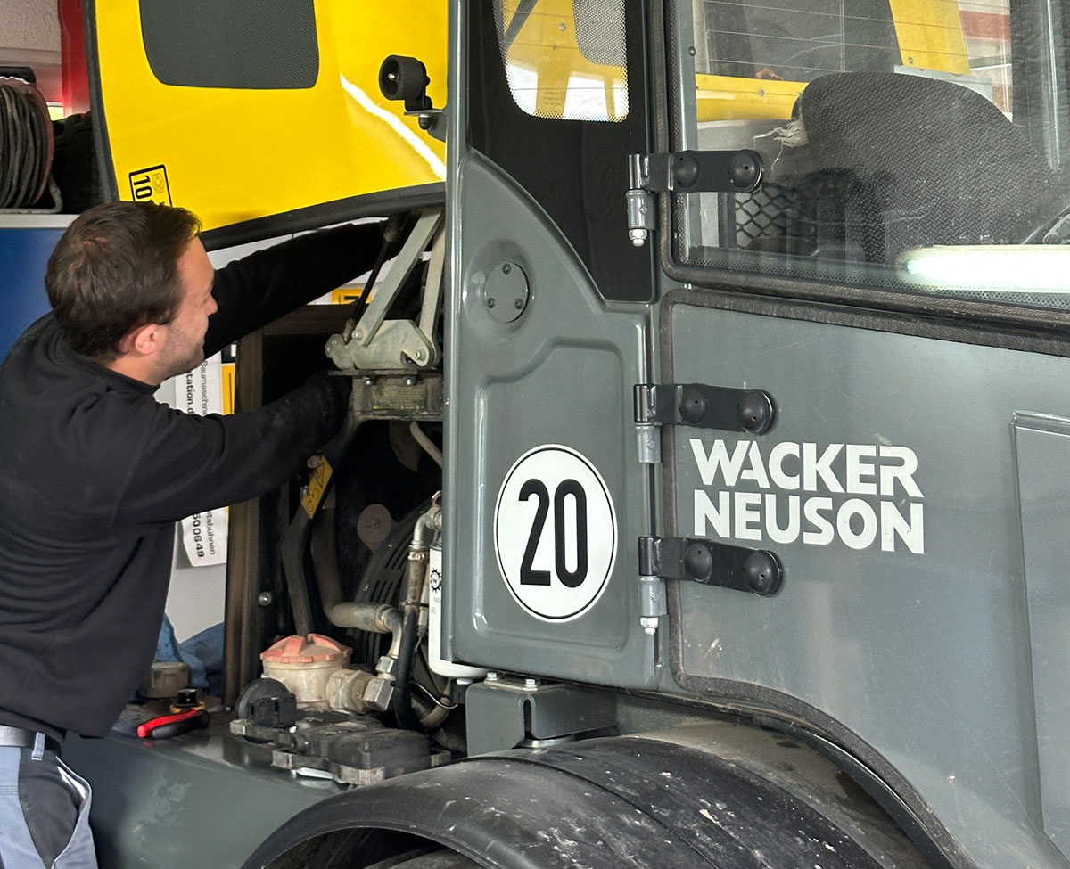 Ein Mann in dunkler Arbeitskleidung repariert den Motorbereich eines grauen Baufahrzeugs mit gelber Motorhaube. An der Fahrzeugtür sind die Aufschrift „WACKER NEUSON“ und die Nummer „20“ in einem weißen Kreis sichtbar.